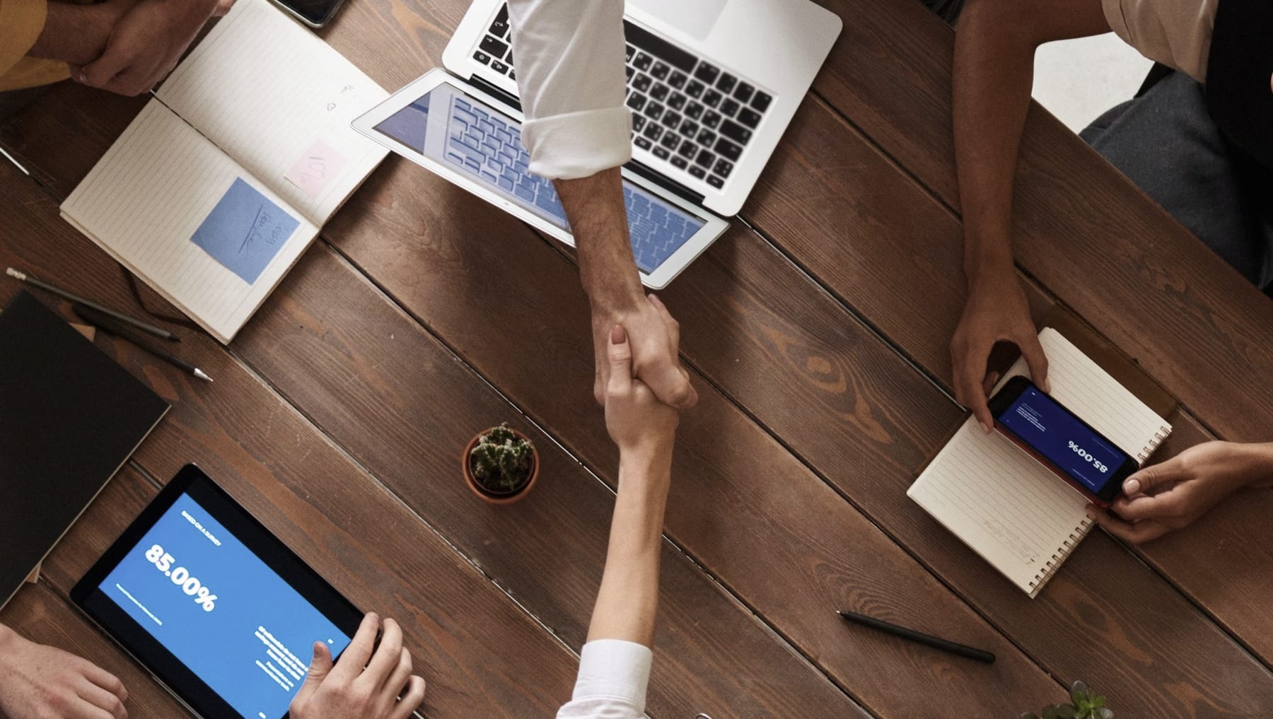 A handshake across a meeting table of open laptops and notebooks.